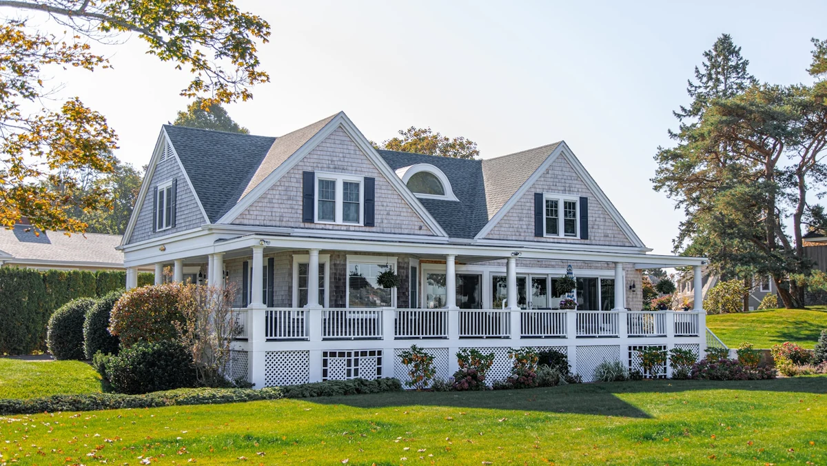 Two-story shingle-style suburban house with wraparound porch on a sunny lawn