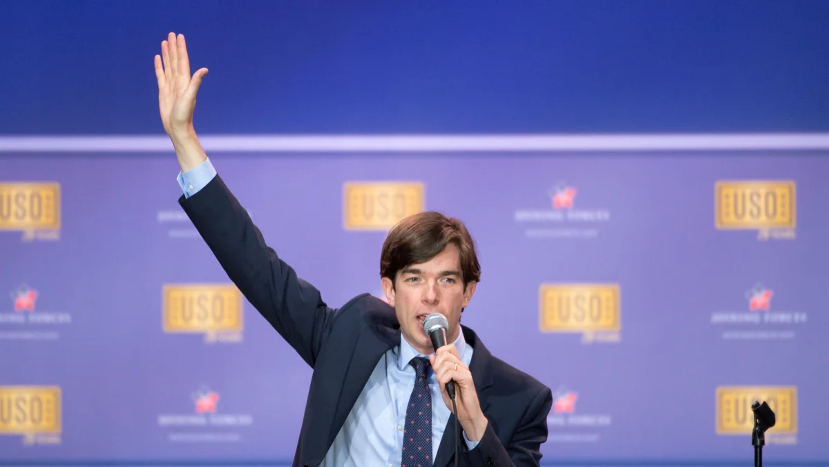 John Mulaney raises one arm and holds a microphone while performing stand-up at a 2016 USO show, years before his Mister Whatever tour