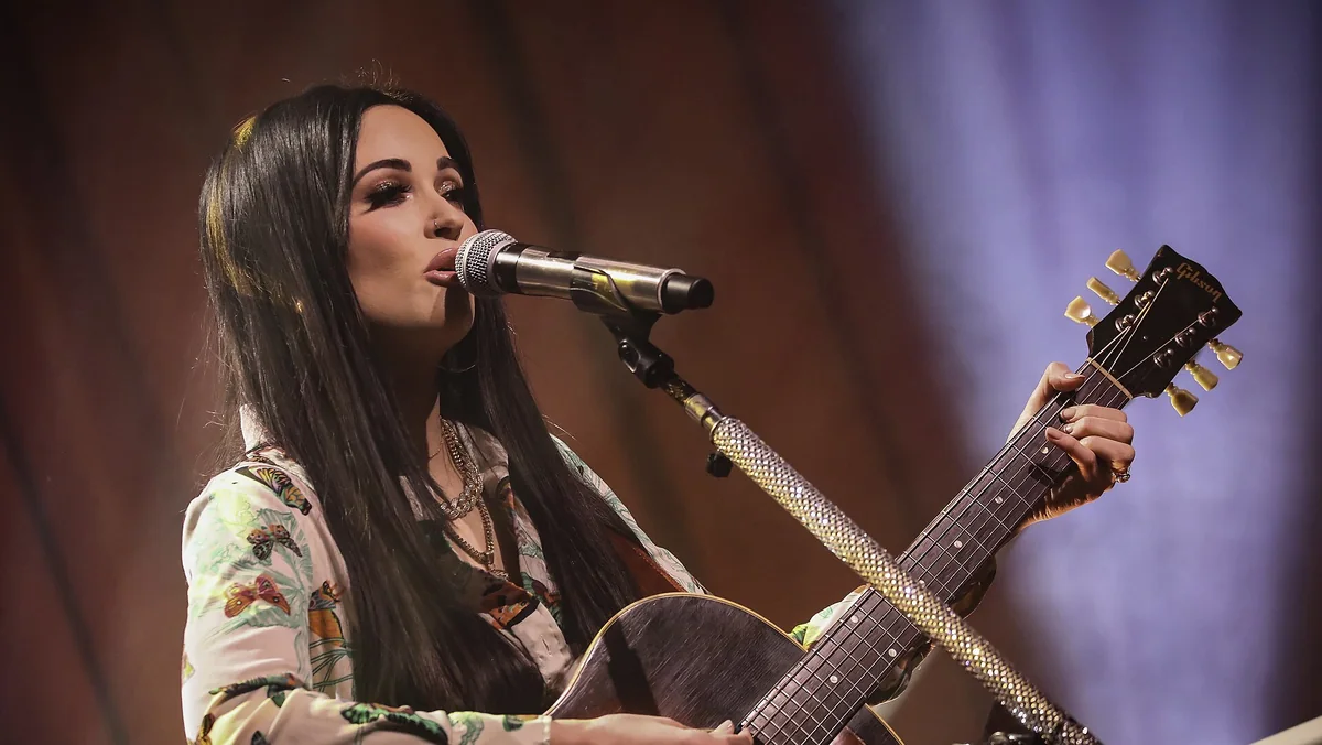 Kacey Musgraves sings into a microphone and plays acoustic guitar under warm stage lights at a 2019 concert in St. Paul, ahead of her Middle of Nowhere Tour stop in Dallas